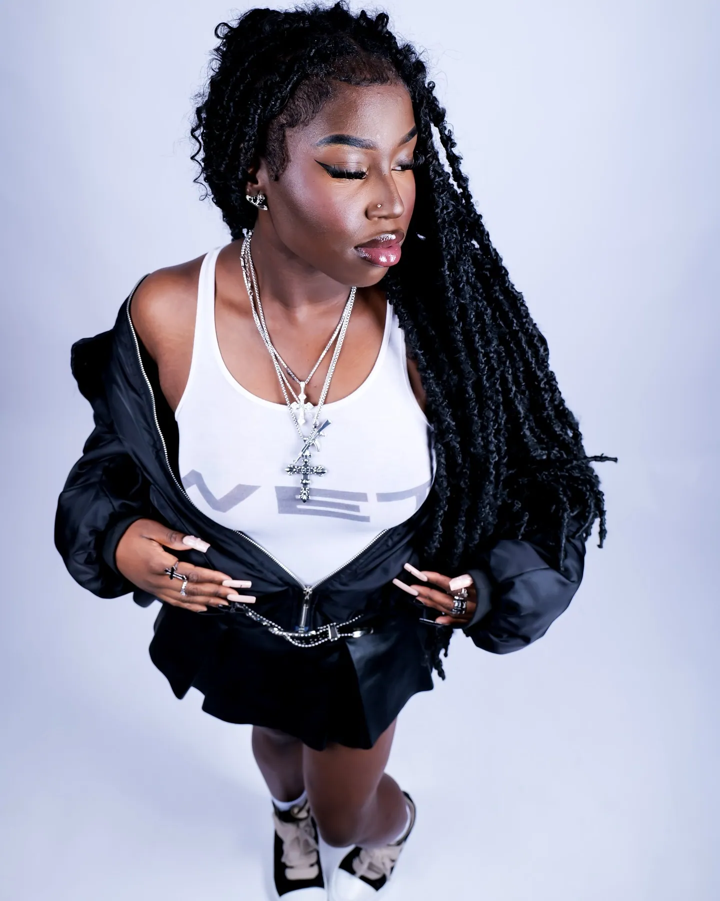 A woman with long braids in a white vest, black jacket and layered chains, looking down at the camera, grey studio backdrop