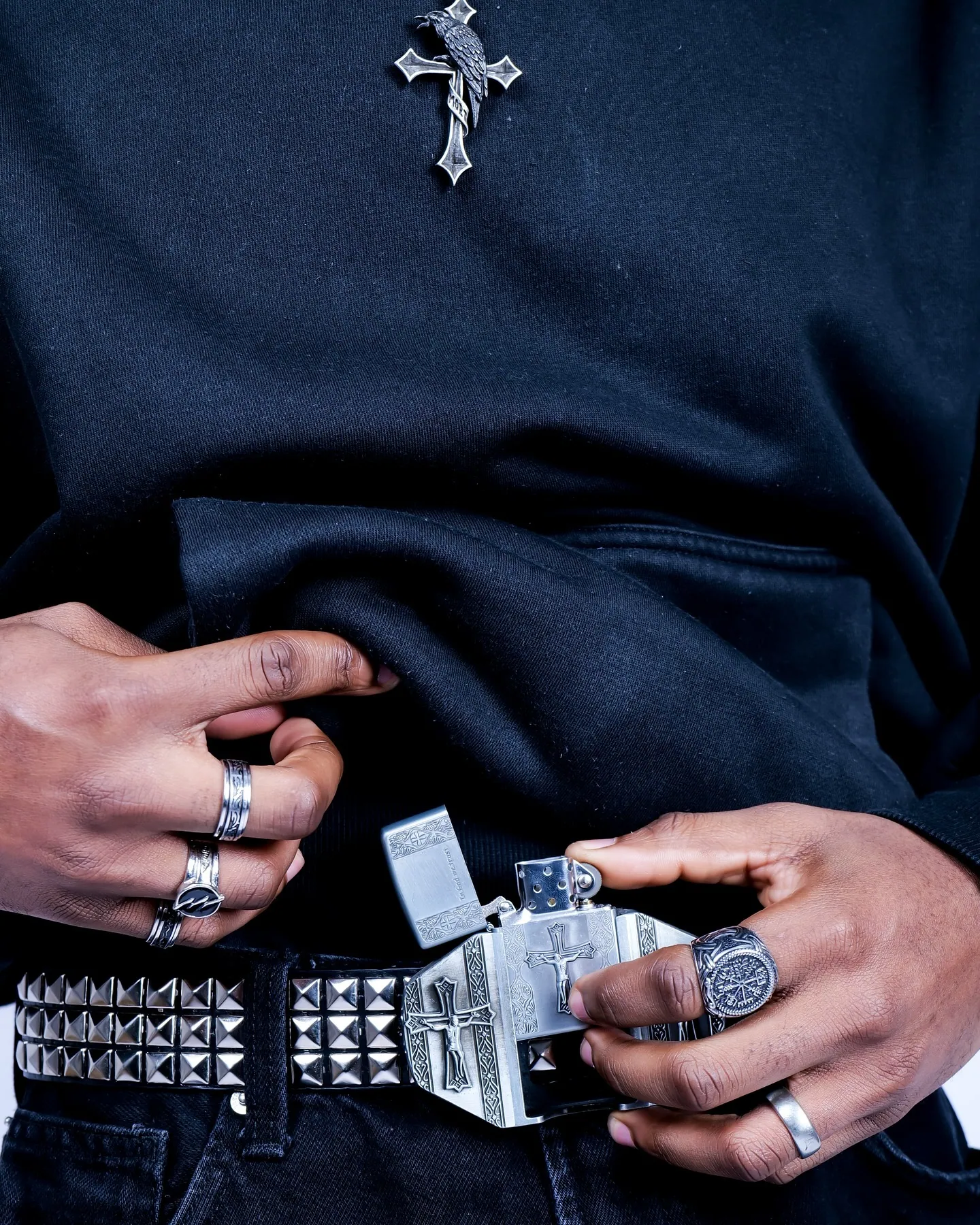 Close-up of a midsection in black clothing with a studded belt, silver rings and a cross pendant, hands on the buckle