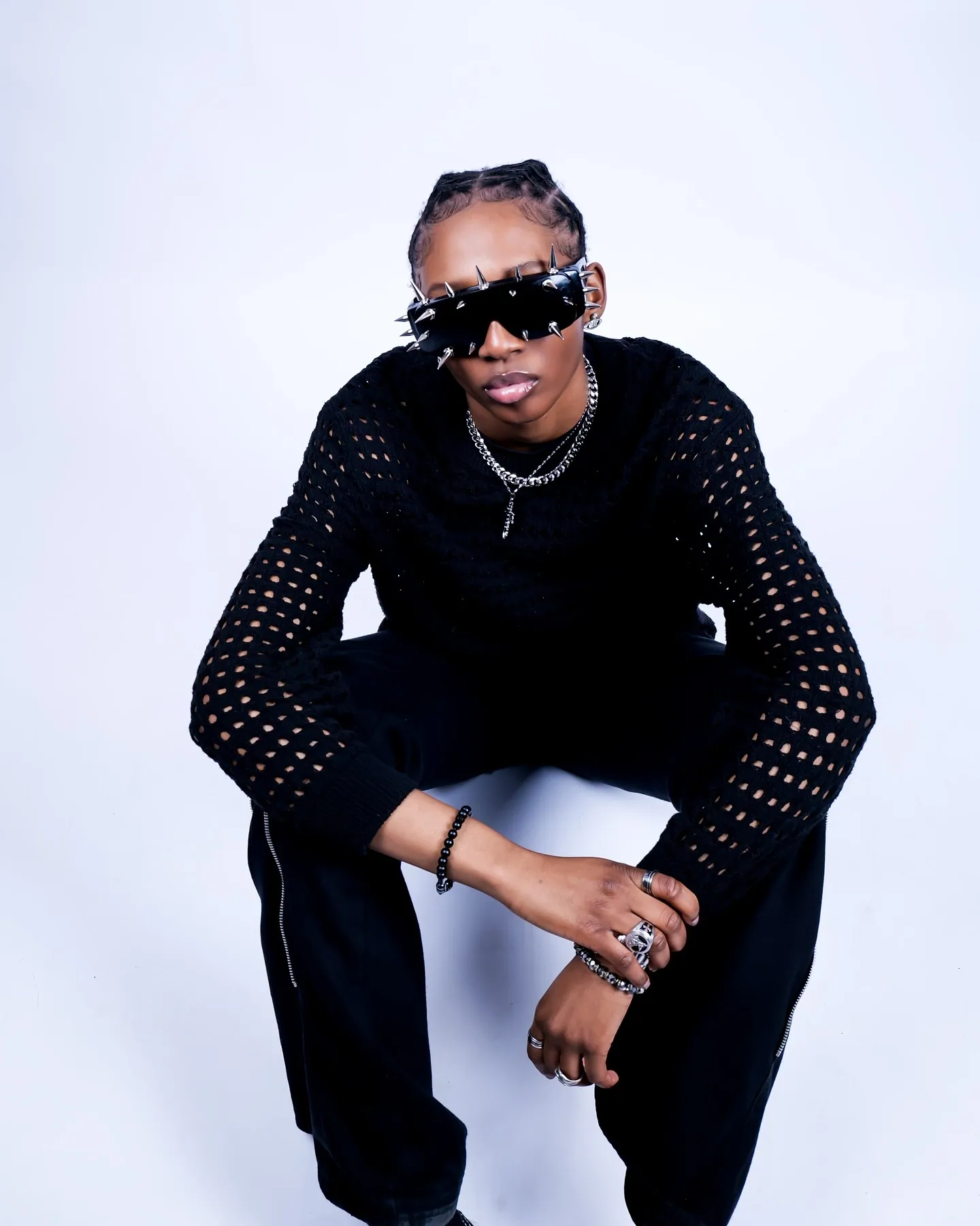 A model crouching in a black mesh-sleeved top, spiked sunglasses and chain necklaces, against a pale backdrop