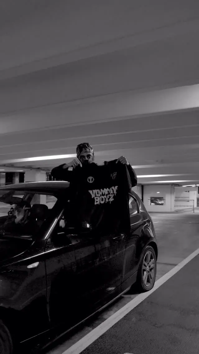 Video still: a man in a car park holding up a black VOMMY BOYZ t-shirt beside a parked car, black and white