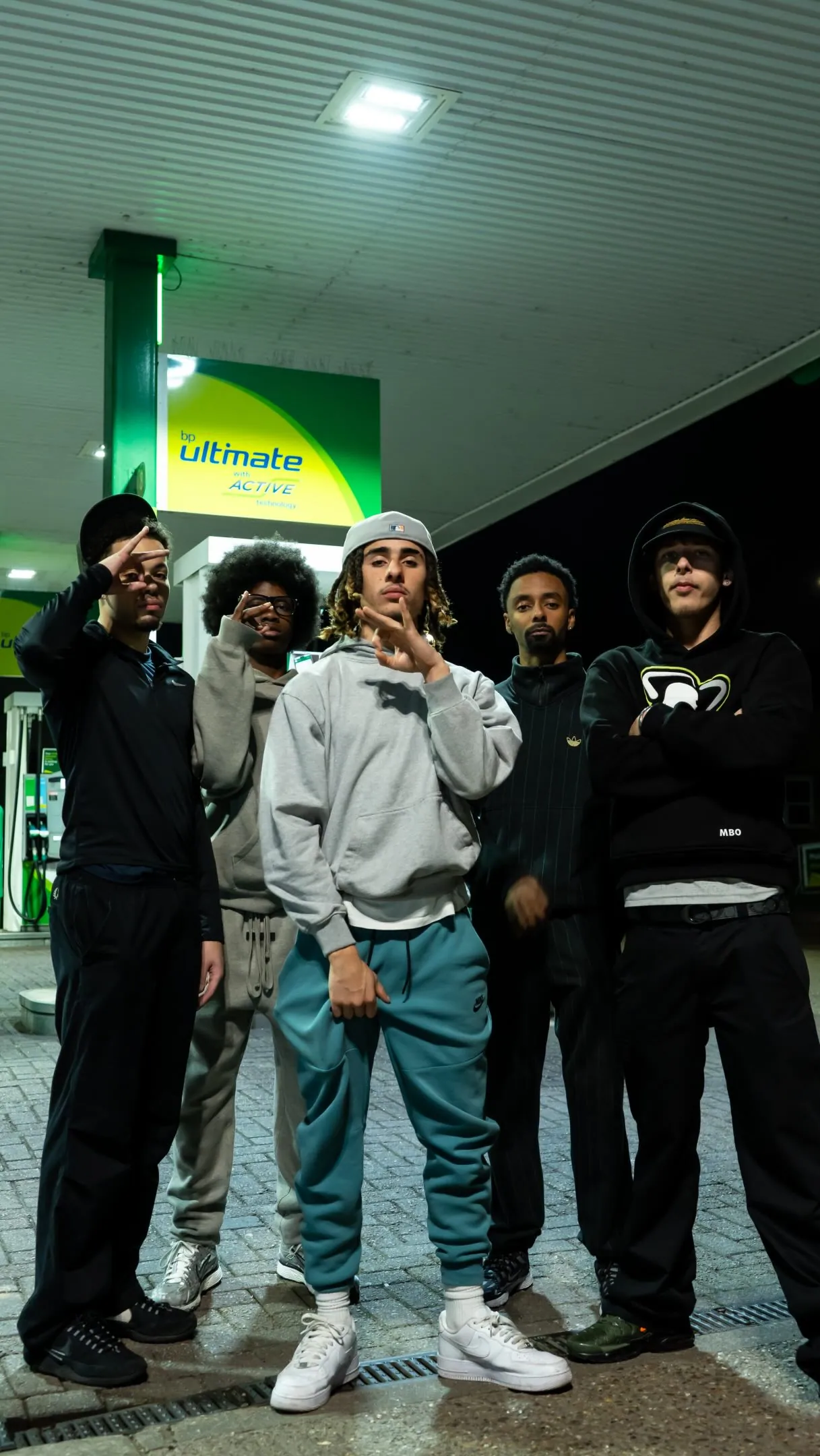 Video still: five young men in dark and grey hoodies and tracksuits posing at night under a BP Ultimate petrol station canopy
