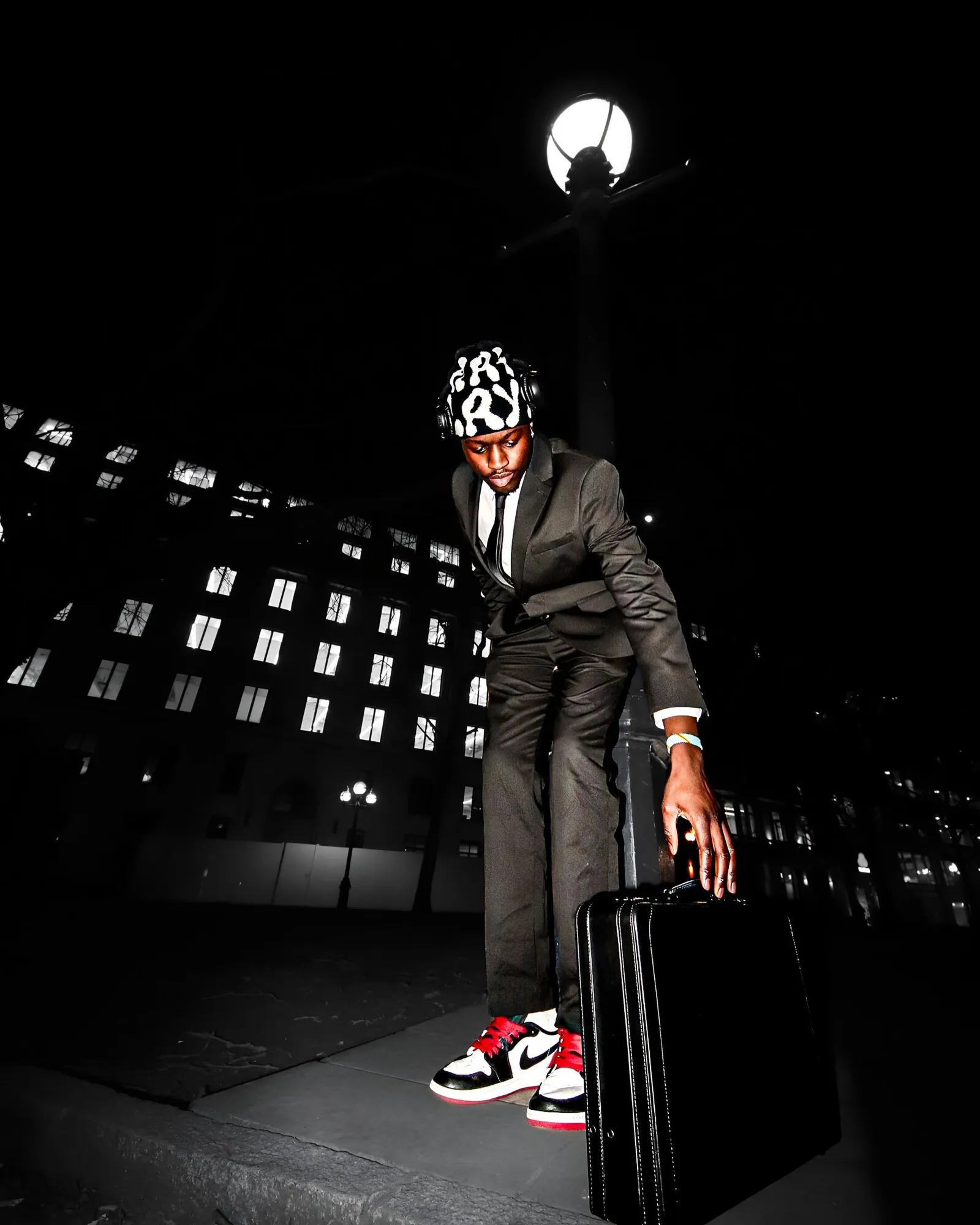 A man in a suit, tie, headphones, patterned beanie and red and white trainers reaching for a briefcase by a night streetlamp