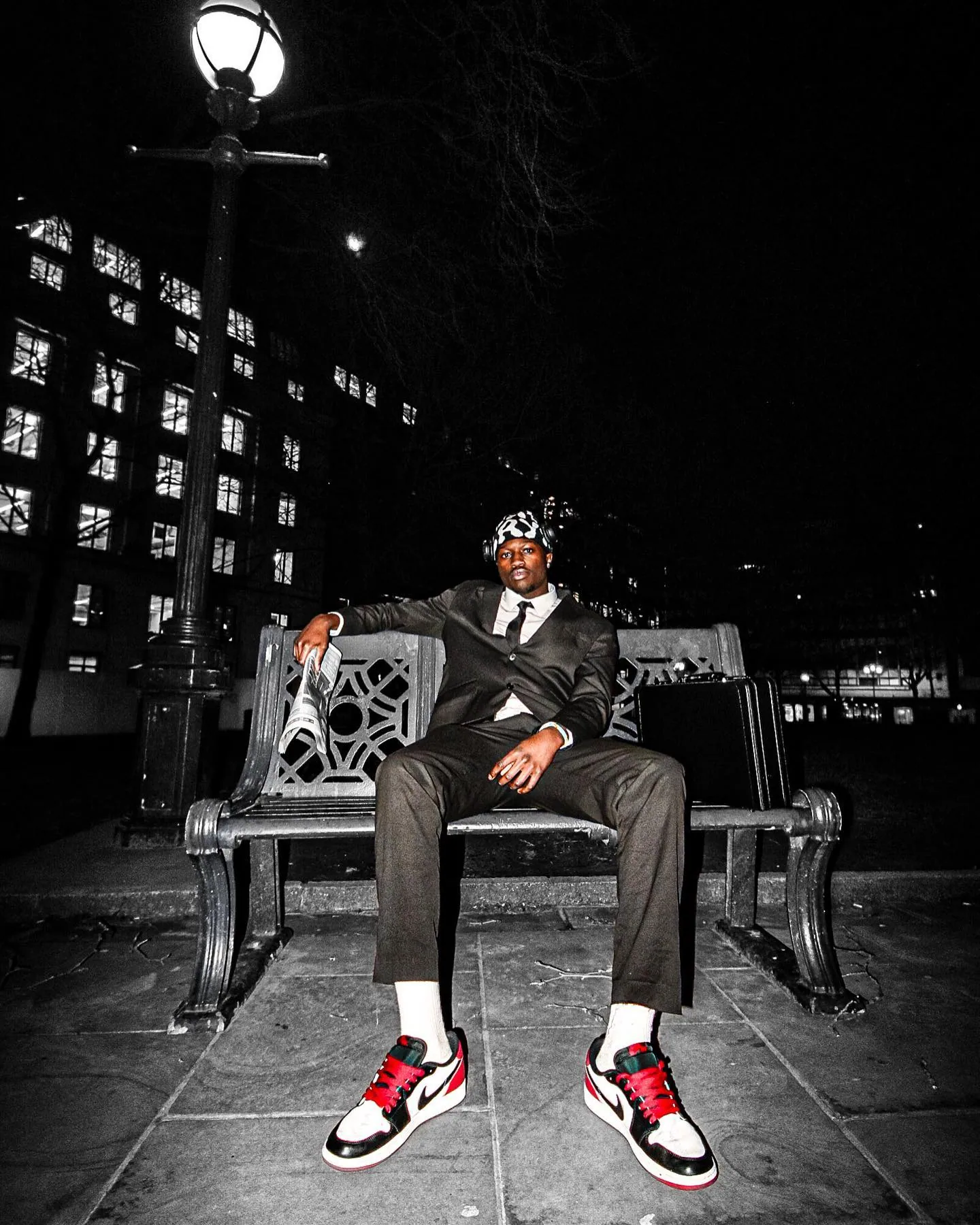 A man in a suit and trainers sitting on an ornate park bench at night with a briefcase and newspaper, streetlamp behind