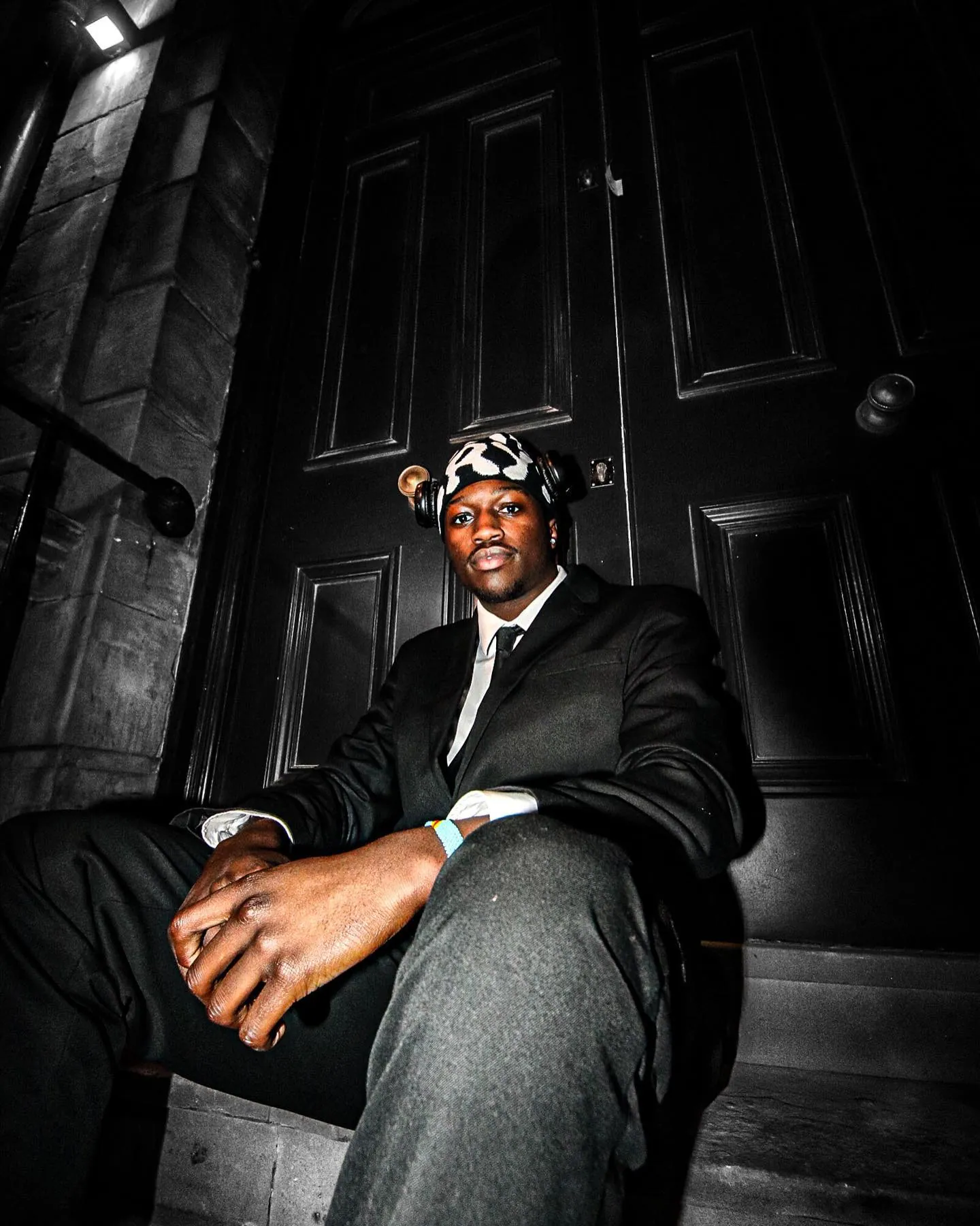 A man in a suit, tie and patterned beanie with headphones sitting on dark steps in front of a black panelled door