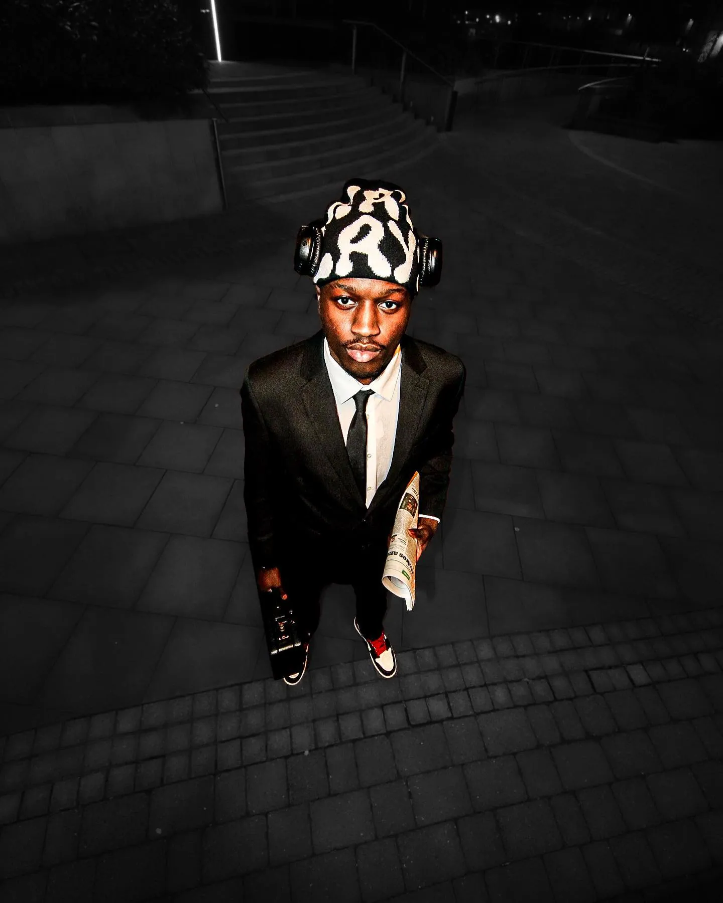 High-angle night shot of a man in a suit, tie and patterned beanie holding a newspaper, standing on dark paving