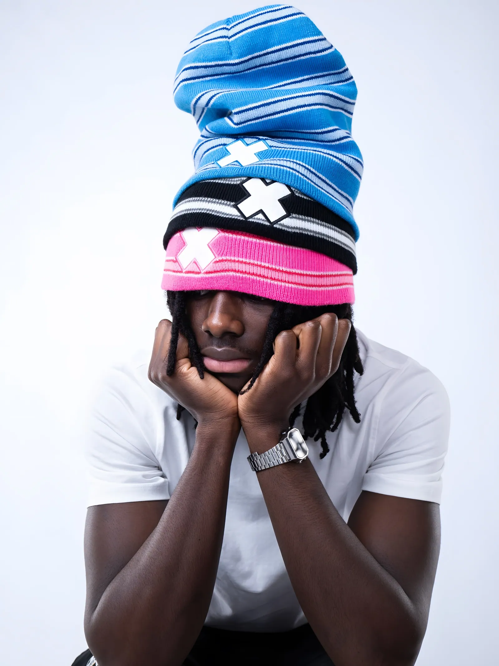 A man resting his face in his hands under a tall stack of blue, black and pink striped beanies with white X patches
