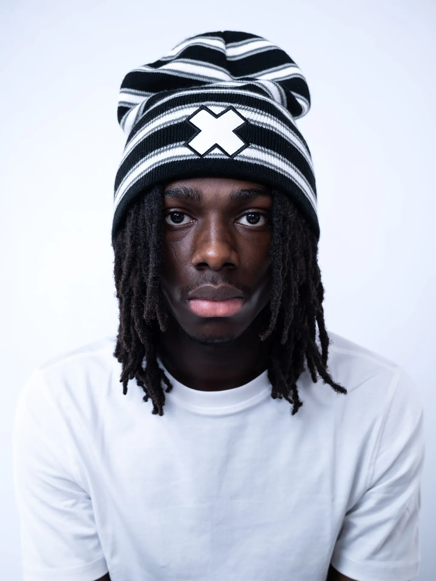 A man with locs in a black and white striped X-patch beanie and white tee, facing the camera against a pale backdrop