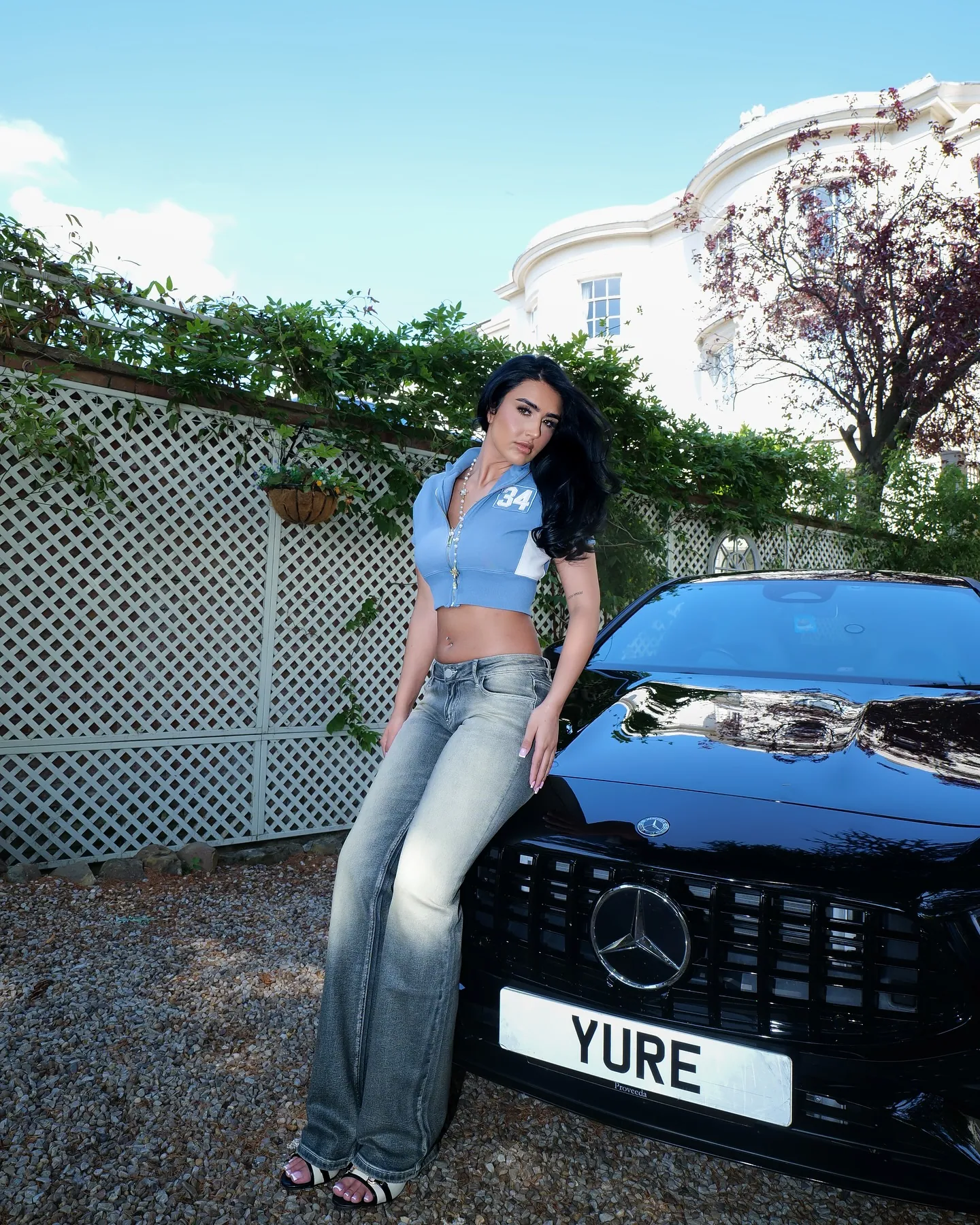 A woman in a blue zip crop top and flared jeans leaning on a black Mercedes with a YURE plate, white lattice fence behind