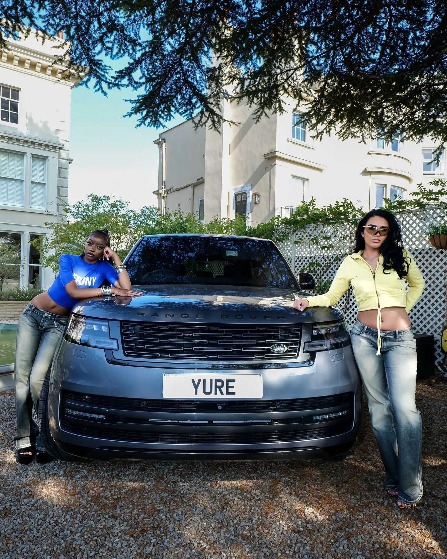 Two women posing against a grey Range Rover with a YURE plate, one in a blue top, one in a yellow cardigan, in a garden
