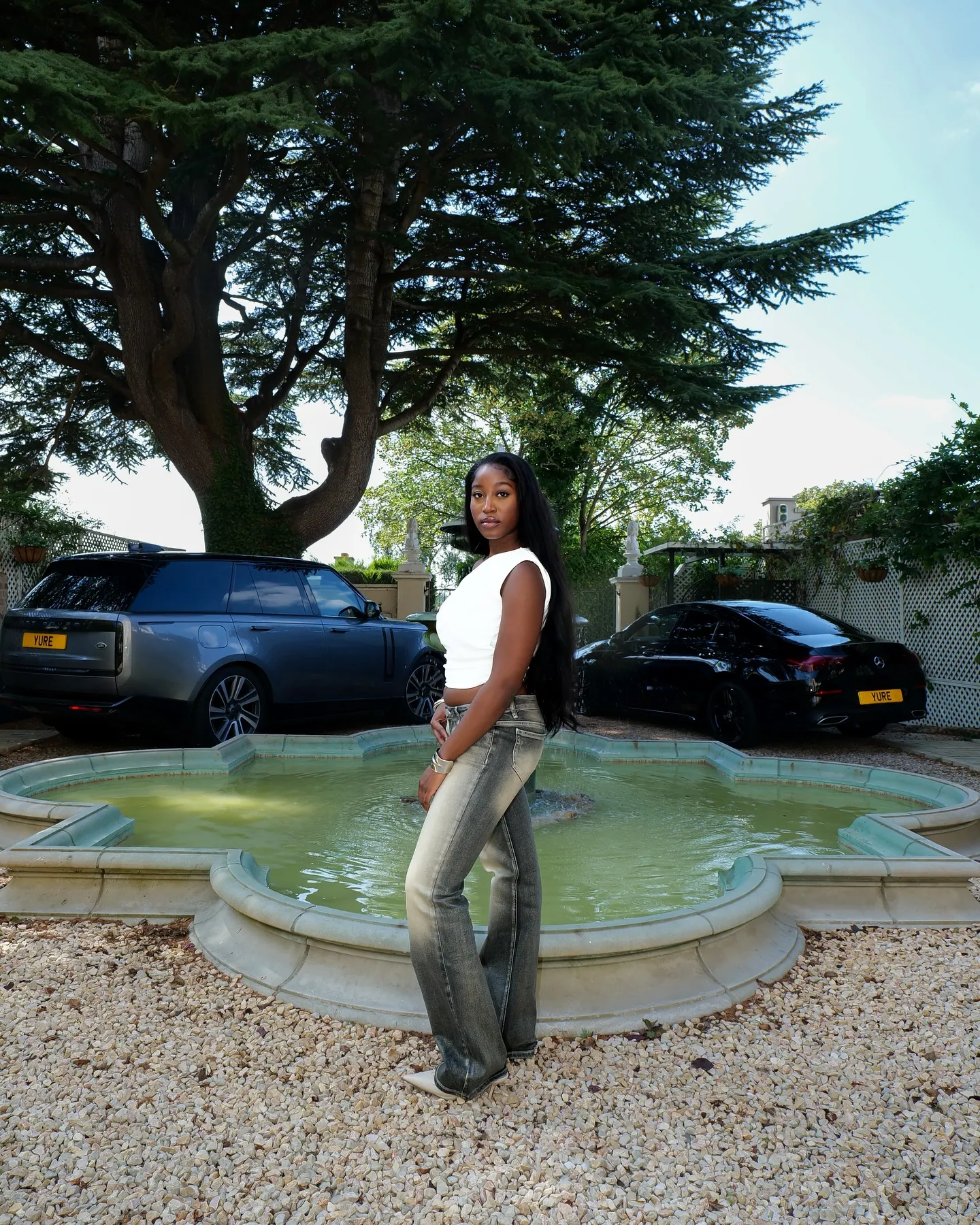 A woman in a white top and flared jeans standing by an ornamental fountain with parked cars and a large cedar tree behind